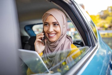 Genç, güzel Müslüman bir kadın elinde tabletle arabanın arka koltuğunda oturuyor ve akıllı telefonuyla konuşuyor. Elinde akıllı telefon tutan ve arabanın arka koltuğunda konuşan gülümseyen genç bir kadın..
