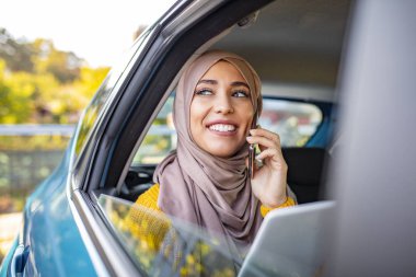 Genç, güzel Müslüman bir kadın elinde tabletle arabanın arka koltuğunda oturuyor ve akıllı telefonuyla konuşuyor. Elinde akıllı telefon tutan ve arabanın arka koltuğunda konuşan gülümseyen genç bir kadın..