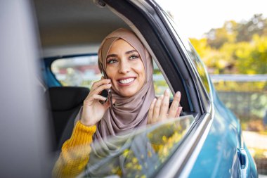 Genç Müslüman iş kadını, arabanın arka koltuğunda telefonla konuşuyor. Güzel Müslüman kadın cep telefonuyla konuşuyor, dijital tablet kullanıyor ve arabada arka koltukta otururken gülümsüyor.