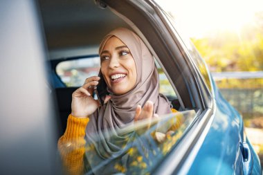 Genç Müslüman bir iş kadınının arabada seyahat ederken cep telefonu kullanması. Bir arabanın arka koltuğunda pencereden dışarı bakan genç bir kadın. Genç Müslüman iş kadını arabada oturuyor..