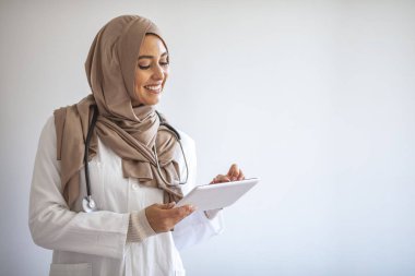 Steteskoplu Muslimah doktor gri arka planda izole edilmiş bir tablet bilgisayar tutuyor. Kendine güvenen Müslüman tıp öğrencisi hastanede poz veriyor. Hastanenin içinde kendine güvenen Müslüman kadın doktor