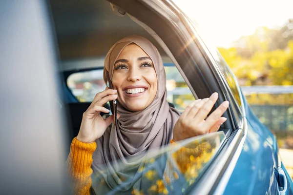 Genç Müslüman iş kadını, arabanın arka koltuğunda telefonla konuşuyor. Güzel Müslüman kadın cep telefonuyla konuşuyor, dijital tablet kullanıyor ve arabada arka koltukta otururken gülümsüyor.
