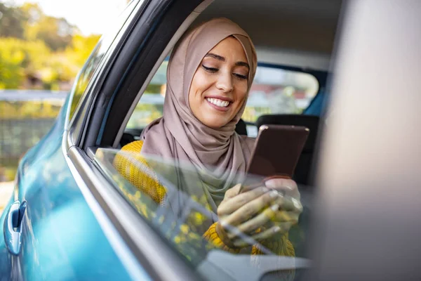 Muslim business woman in car. Writing a message on her mobile phone. Positive pensive Islamic woman in hijab sitting on backseat of taxi and drinking coffee while using gadget