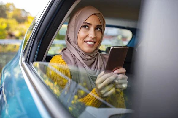 Portrait of happy beautiful young Muslim woman in beige hijab sitting on backseat of car. Beautiful Muslim business woman is using a smart phone and smiling while sitting on back seat in the car
