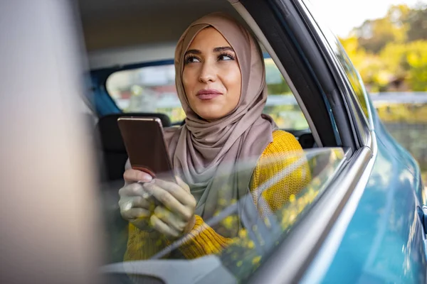 Portrait of happy beautiful young Muslim woman in beige hijab sitting on backseat of car. Beautiful Muslim business woman is using a smart phone and smiling while sitting on back seat in the car