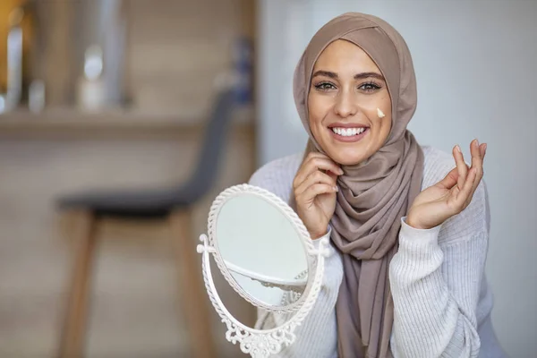 Muslim woman spreading cream over her face while looking in the mirror. Beauty treatment. Female putting on moisturizer on her facial. Muslim woman applying cream to face and looking to mirror