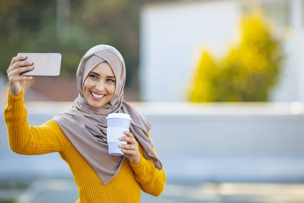 若いイスラム教徒の女性が自撮りを手に飲み物を持っています 携帯電話でヒジャーブの幸せなアラブ女性が自撮りをしています 笑顔の少女の肖像 屋外に行くためにコーヒーを振り — ストック写真