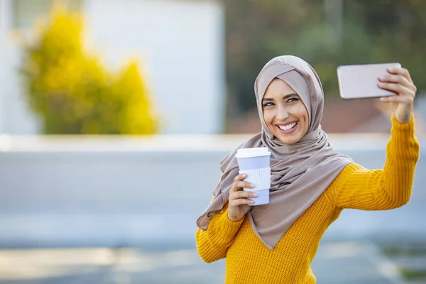 若いイスラム教徒の女性が自撮りを手に飲み物を持っています 携帯電話でヒジャーブの幸せなアラブ女性が自撮りをしています 笑顔の少女の肖像 屋外に行くためにコーヒーを振り — ストック写真