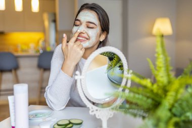 Evde yüzünde temizlik maskesi olan genç bir kadın. Cilt bakımı. Güzellik maskesi takan kadın, yaklaşın. Çok güzel. Yüzünde güzellik maskesi olan ve aynaya bakan kızın yakın çekimi..