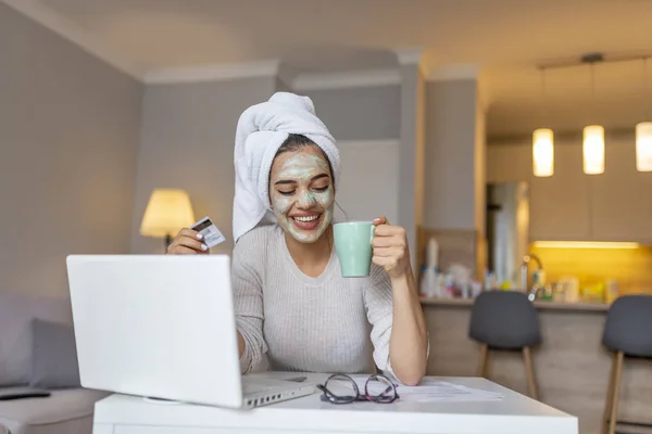 Woman with cosmetic face mask shopping online from her home. Leisure time at home. pPortrait of beautiful woman with facial taking a coffee at home while use internet with modern laptop computer