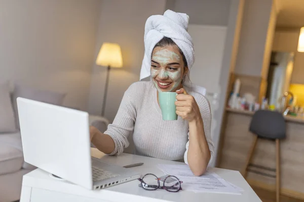 Young woman with cosmetic mask on face working with laptop at home in the evening. Girl is working from home, she has face mask and she is in the bath robe.