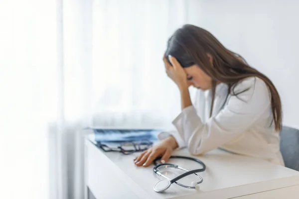 Stethoscope and doctor sitting with laptop stress headache about work in hospital. Frustration young female doctor sitting in her consulting room and looking at document.