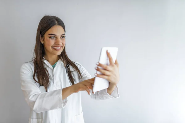 Doctor with digital tablet. Attractive young female doctor in white lab coat working on digital tablet and smiling while standing against grey background.