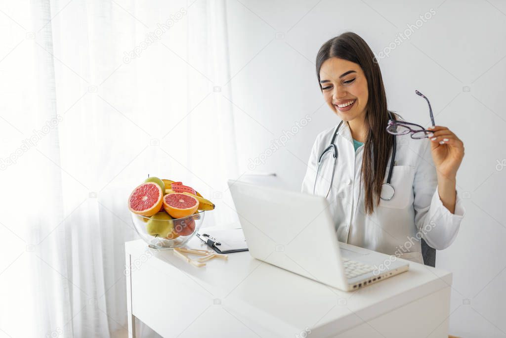 Nutritionist doctor with fruits in office. Nutritionist working on laptop and writing diet plan for patient. Portrait of female nutritionist in her office. Smiling young dietician sitting at desk 