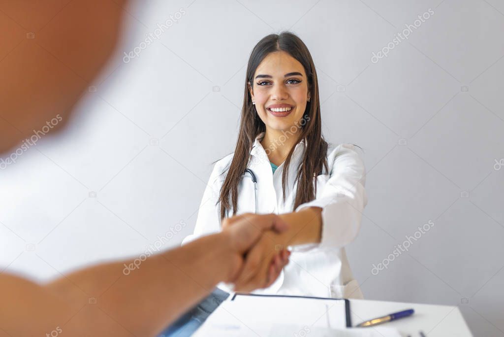 Female doctor in white coat smiling while shaking hand to male Patient blurred in foreground. Partnership, trust and medical ethics concept. Female doctor handshaking a patient's hand and smiling