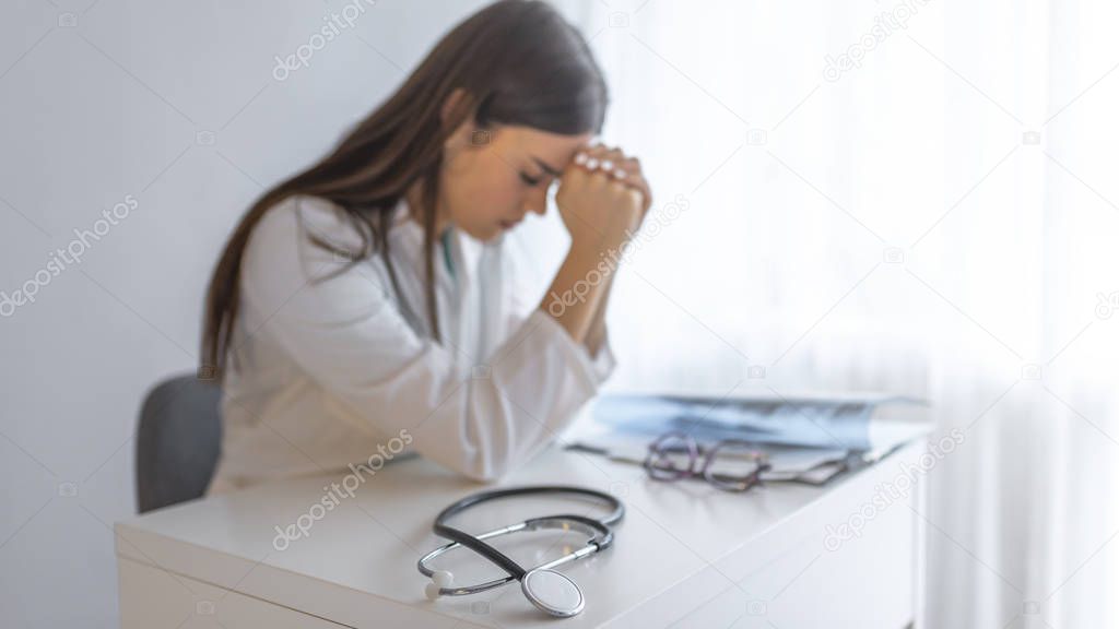 Close up stethoscope and doctor sitting with laptop with stress headache. Stressed female doctor sat at his desk. Mid adult female doctor working long hours.