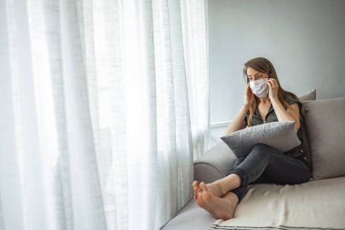 Genç bir kadın soğuk ve kanepede dinleniyor. Tıbbi maskeli kadın evde karantina altında kalıyor. Yüz maskesi takmış karantinaya alınmış bir kadın. Evde pencerenin önünde tek başına oturuyor.