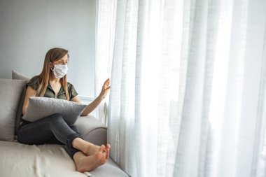 Tıbbi maskeli genç bir kadın kendini karantinaya almak için evde karantinada kalıyor. Ev karantinası, COVID-19 önleme, Coronavirüs salgını durumu. Coronavirüs salgını