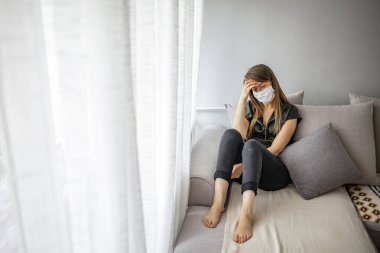 Tıbbi maskeli genç bir kadın kendini karantinaya almak için evde karantinada kalıyor. Ev karantinası, COVID-19 önleme, Coronavirüs salgını durumu. Coronavirüs salgını