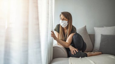 Sad lonely girl isolated stay at home in protective sterile medical mask on face looking at window, bored woman because of Chinese pandemic coronavirus virus covid-19. Quarantine, prevent infection clipart