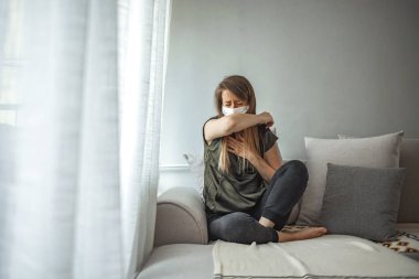 Coronavirus hastalığı (COVID-19) belirtileri - burun akıntısı, boğaz ağrısı, öksürük, ateş. Yüzü maskeli genç bir kadın grip virüsünden hasta. Evde karantina altında yatan Corona virüsü yayıyor.
