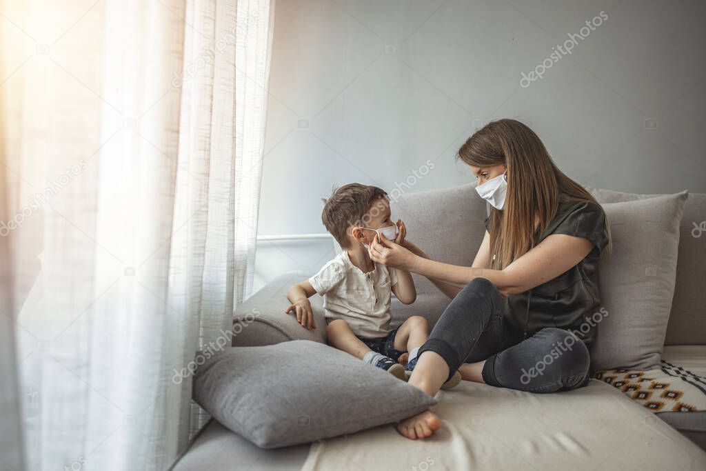 Side view of adult woman putting on medical mask on little boy during coronavirus outbreak against gray background. Mother and her son putting masks on protect themselves from viruses