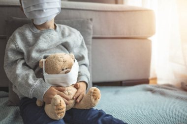 Evde karantinada oturan çocuk, hasta oyuncak ayısıyla Coronavirus ve grip salgını sırasında virüslere karşı maske takıyor. Çocuklar ve hastalık COVID-2019 hastalık konsepti
