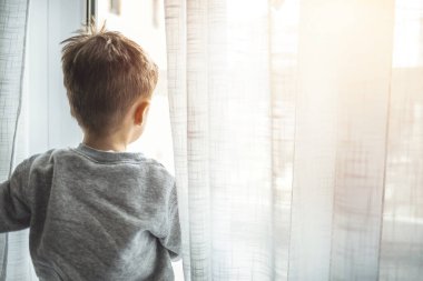 Çocuk karantinada. Çocuk pencere pervazına oturur ve pencereden dışarı bakar, evde oturur, yürümek ister. Virüs koruması, koronavirüs salgını, önleme salgını. Koronavirüs salgınını önlemek için karantinada kalın..