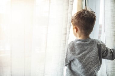 Çocuk karantinada. Çocuk pencere pervazına oturur ve pencereden dışarı bakar, evde oturur, yürümek ister. Virüs koruması, koronavirüs salgını, önleme salgını. Koronavirüs salgınını önlemek için karantinada kalın..