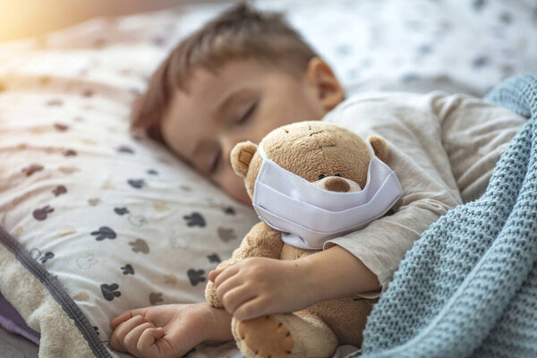 Little boy sleeping in his bed with his Teddy bear wearing a mask to protect him against corona virus covid-19 / 2019-nCov concept. Adorable toddler boy taking a nap in a white bed holding his teddy bear