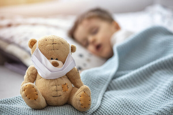 Child in home quarantine at the bed, sleeping, with medical mask on his sick teddy bear, for protection against viruses during coronavirus COVID-19 and flu outbreak. Children and illness disease concept