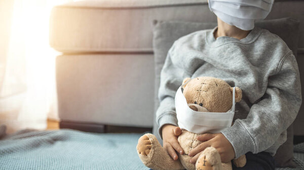 Child in home quarantine at the window putting a medical mask on his sick teddy bear, for protection against viruses during coronavirus COVID-19 and flu outbreak. Children and illness disease concept