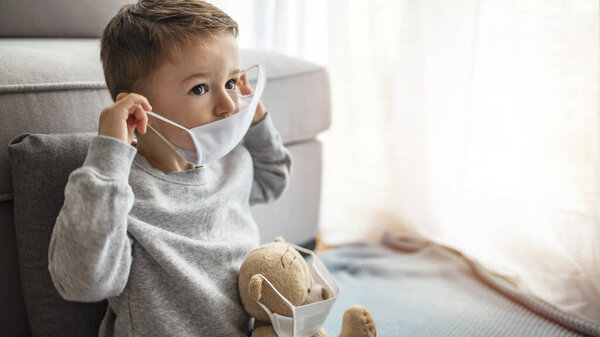 Child in home quarantine looking out of the window with his sick teddy bear wearing a medical mask against viruses during coronavirus and flu outbreak. Children and illness COVID-2019 disease concept