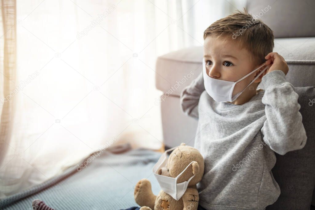 Quarantine, threat of coronavirus, virus protection, pandemic. Child and his teddy bear both in protective masks sits on windowsill inside house and looks out the window. Coronavirus epidemic. 