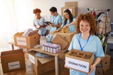 Happy volunteer looking at camera. Woman organizes food and clothing drive. She is smiling at the camera and is holding a donation box. Small group of people working on humanitarian aid project clipart