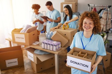 We are successful team of social workers. Woman volunteer holding a donation box and is smiling at the camera. Volunteers are helping people in the background. clipart