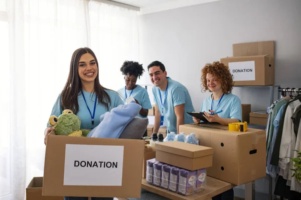 Happy volunteer looking at camera. Woman organizes food and clothing drive. She is smiling at the camera and is holding a donation box. Small group of people working on humanitarian aid project