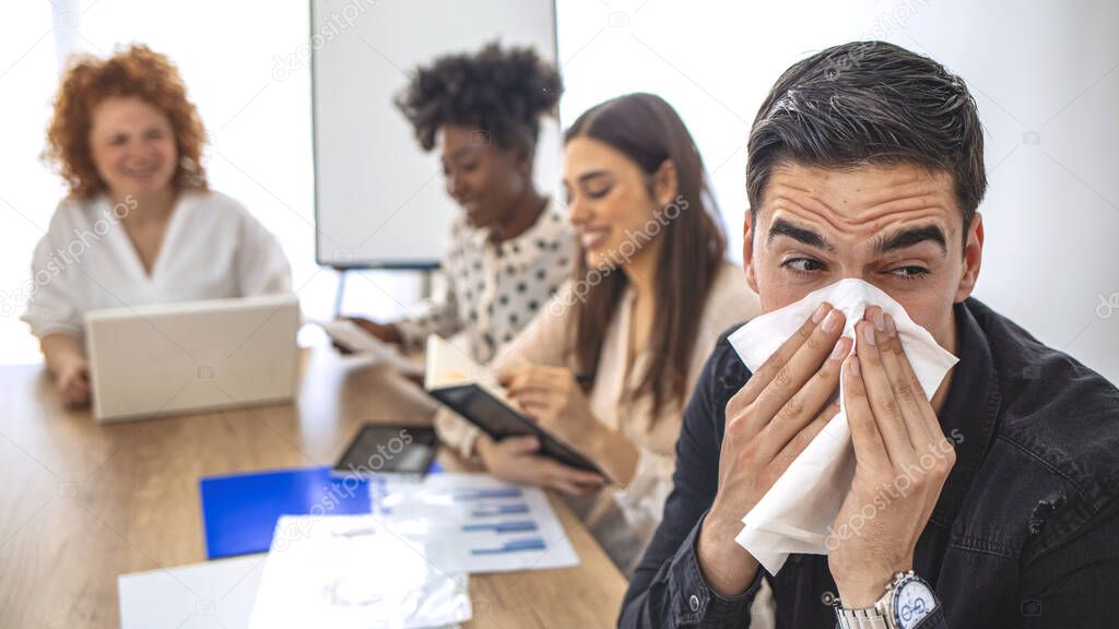 Cropped shot of a businessman blowing his nose while his colleagues look. Cropped shot of a sick businessman blowing her nose in the boardroom. Businessman suffering with allergies in an office