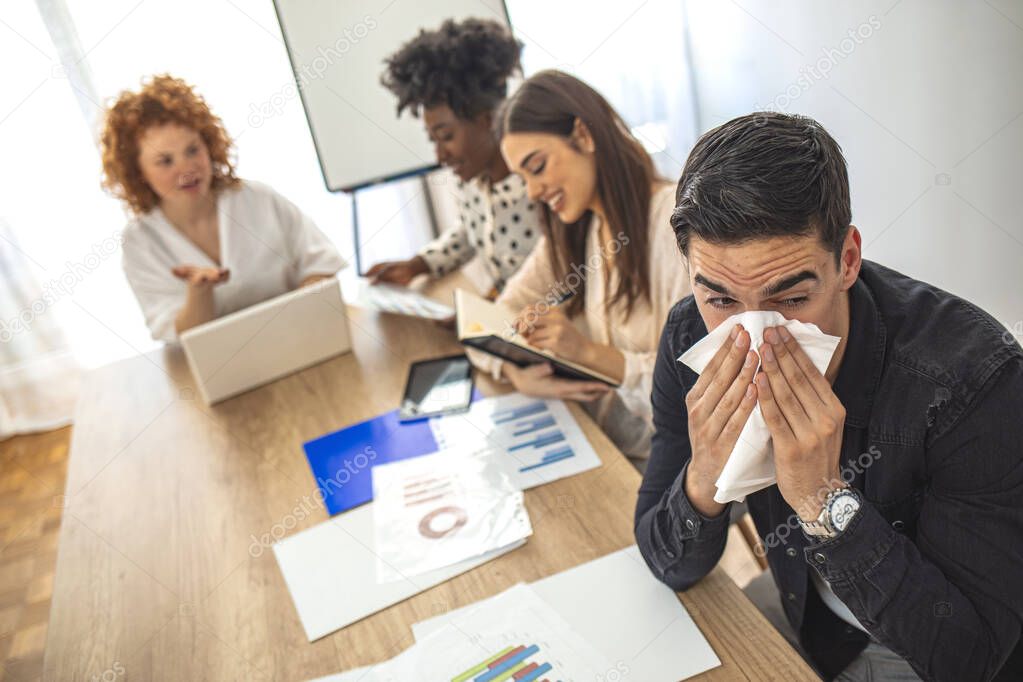 Shot of a frustrated businessman using a tissue to sneeze. Sick man with handkerchief sneezing blowing nose while working with coworkers, businessman caught cold.  Pandemic influenza, disease prevention