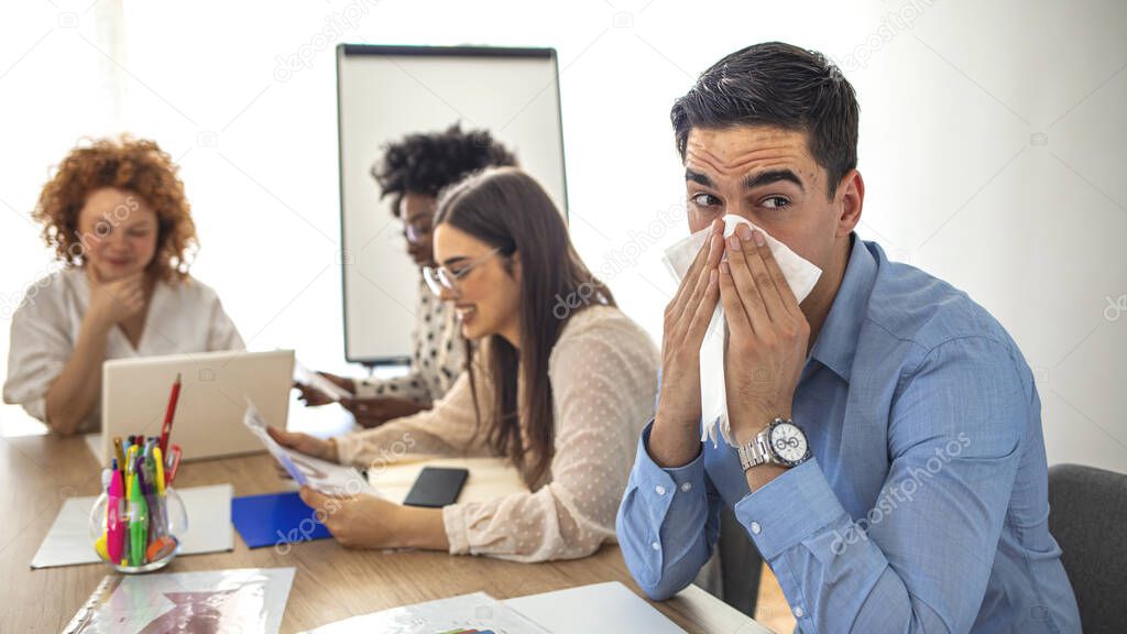 Cropped shot of a businessman blowing his nose while his colleagues look. Cropped shot of a sick businessman blowing her nose in the boardroom. Businessman suffering with allergies in an office