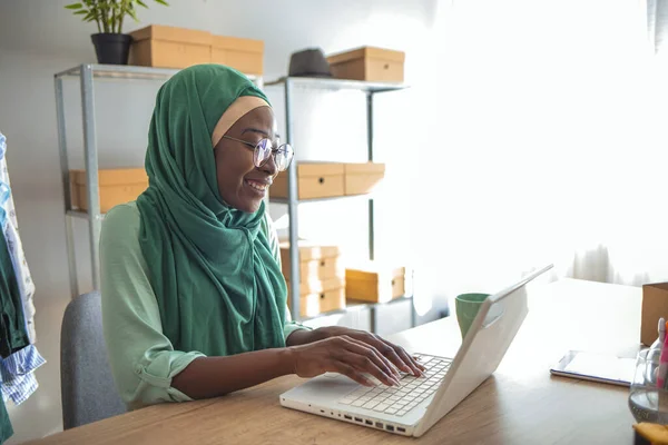若いアラビア人女性起業家は ヒジャーブを着て自宅のテーブルに座り 文書を読み ノートパソコンで作業しています 彼女の仕事を成し遂げるためには — ストック写真