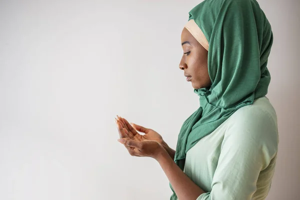 Young muslim woman in green hijab and traditional clothes praying for Allah, copy space. Arab Muslim woman praying, isolated on grey background. Humble Muslim woman holding hands up and praying in peace