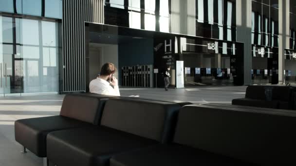 Hombre de negocios hablando por teléfono en un gran centro de negocios modernizado con techos grandes y paredes espejadas, sentado en un sofá de cuero — Vídeo de stock