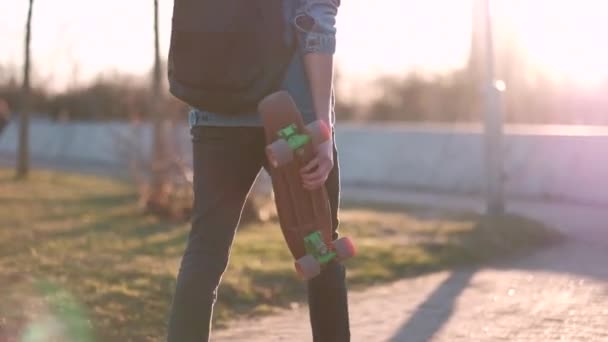 Молодой парень ходит со скейтбордом в руках, на закате, в джинсовой куртке и черных джинсах — стоковое видео