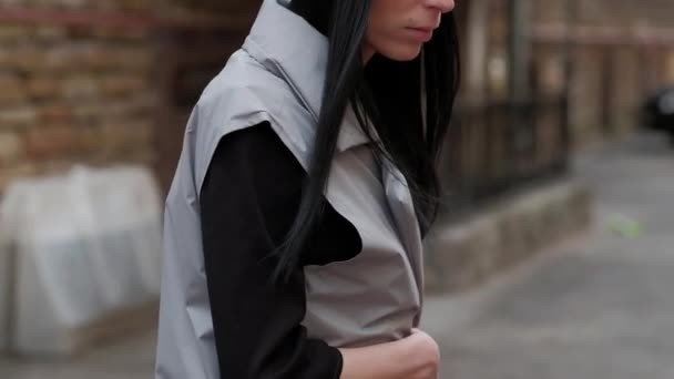 背景のレンガの壁、灰色のマントと黒い帽子に、通りを歩いて、黒い長い髪と、スタイリッシュなファッションの女の子 — ストック動画
