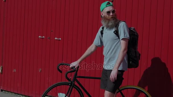 Un hombre adulto, con una gran barba y una elegante bicicleta negra, de pie contra la brillante pared roja, en pantalones cortos, gafas, verde, sombrero y mochila — Vídeos de Stock