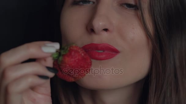 Mädchen mit roten Lippen und ausdrucksstarken blauen Augen schaut in die Kamera und lächelt verschmitzt eine große Erdbeere — Stockvideo