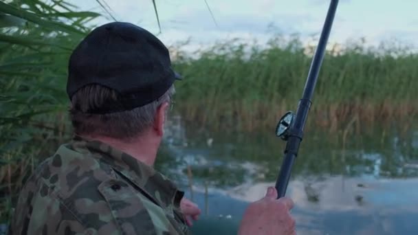Střelba rukou: Rybář háček ryby, muž v maskování ryby v jezeře v rákosí v lodi — Stock video