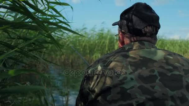 Rybak w kurtka kamuflaż i czarna Czapka siedzi w łodzi czeka na ryby ugryźć — Wideo stockowe