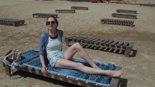Attraktives Mädchen, das sich am Strand ausruht, seinen Freunden zuwinkt, seine Freunde sieht, auf hölzernen Liegestühlen sonnt — Stockvideo